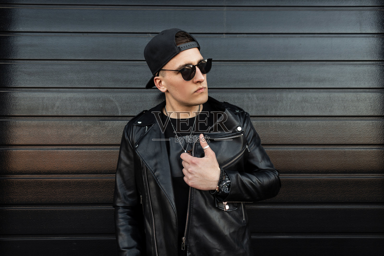 时尚的年轻嬉皮士男士，戴着时髦的深色太阳镜，穿着黑色皮夹克和时尚的棒球帽，正在一座现代黑色木屋旁摆姿势。帅气的男孩在街上，复古风格。照片摄影图片