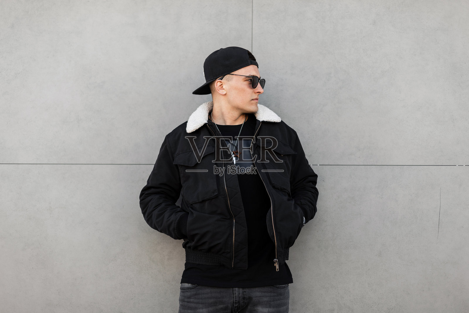 年轻的现代时尚男士，戴着黑色棒球帽，佩戴时尚的深色太阳镜，穿着带白色毛皮的时尚黑色夹克，搭配灰色牛仔裤，站在一座复古建筑旁。英俊的城市男模在散步。照片摄影图片