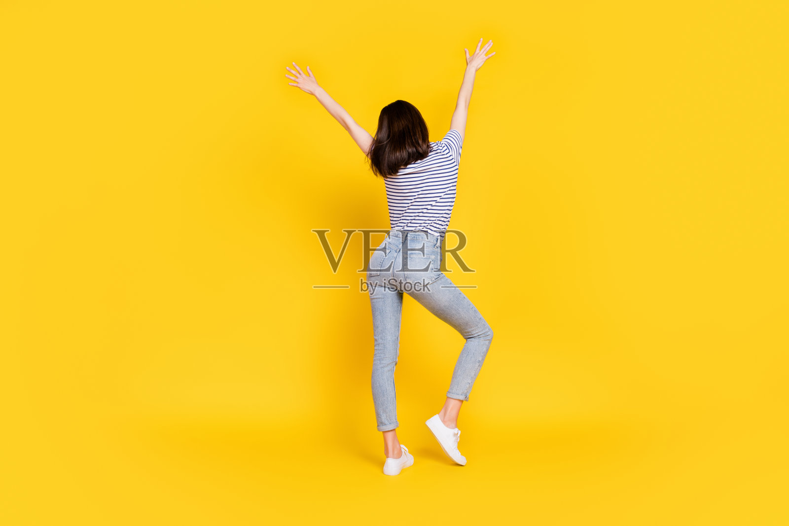 背影肖像的漂亮女士，穿着时尚的牛仔裤和条纹T恤，抬起手臂，背景为黄色。照片摄影图片
