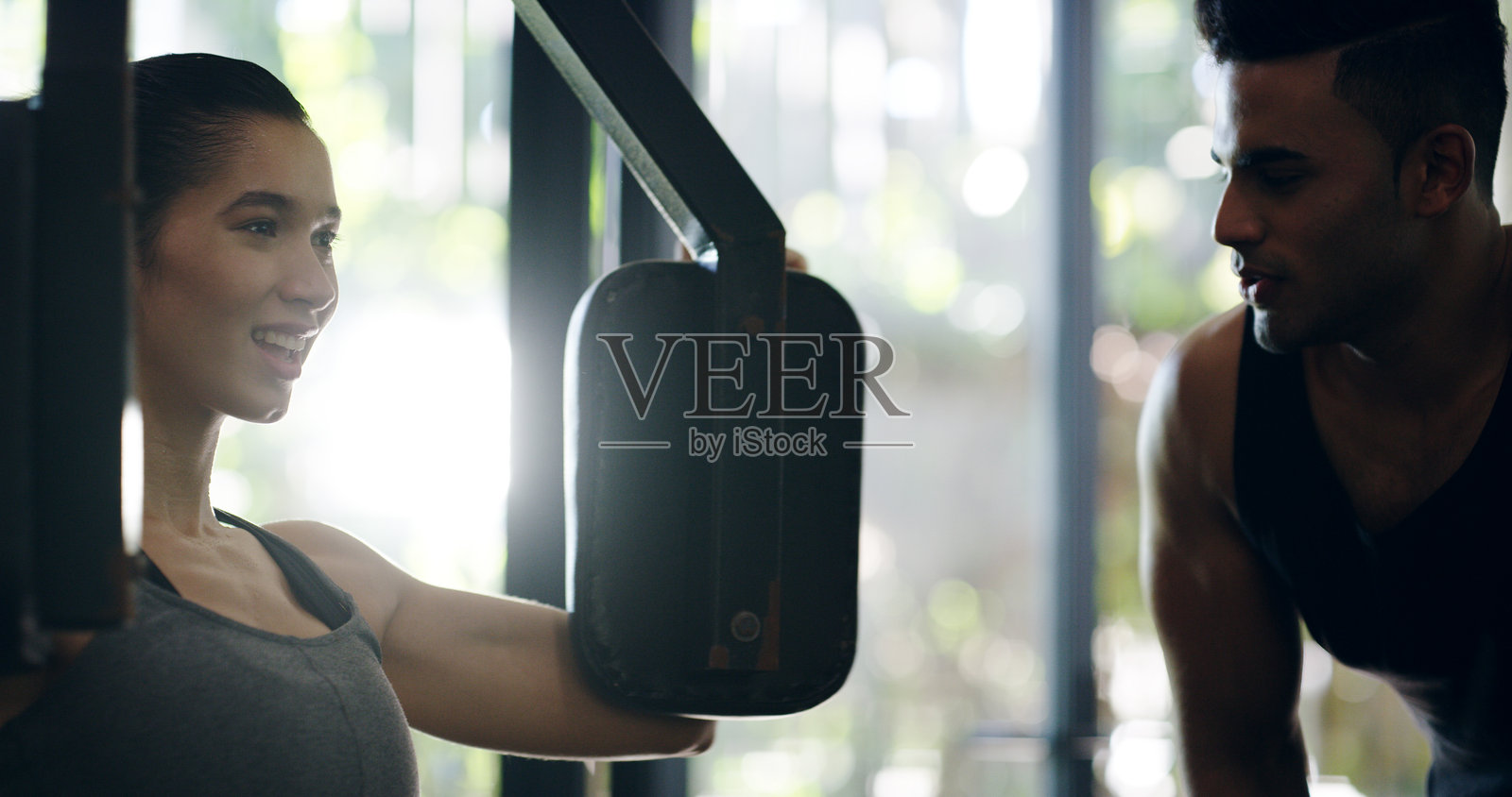 健身房中与女性一起进行胸部锻炼的器械和私人教练，旨在提升身体素质或力量训练。充满力量、微笑和自信的人在器械上挑战自我，进行健身锻炼。照片摄影图片