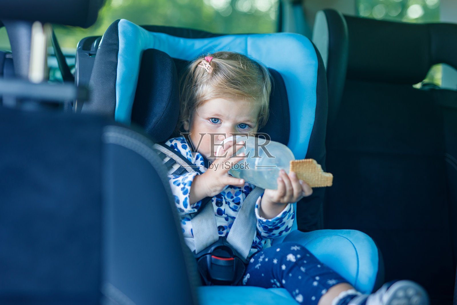 可爱的蓝眼睛小女孩坐在汽车安全座椅上。幼儿正在进行家庭旅行和旅程。在交通堵塞中，快乐地微笑，喝着奶瓶里的牛奶，吃着饼干。照片摄影图片