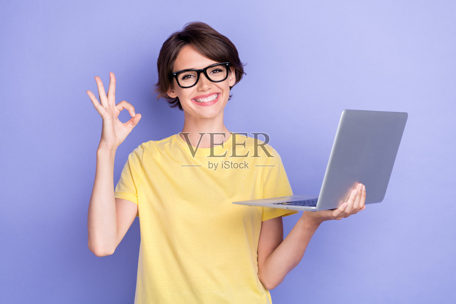 年轻迷人的可爱女性穿着黄色T恤，手持笔记本电脑，露出OK手势和灿烂的微笑，背景为紫色。照片摄影图片