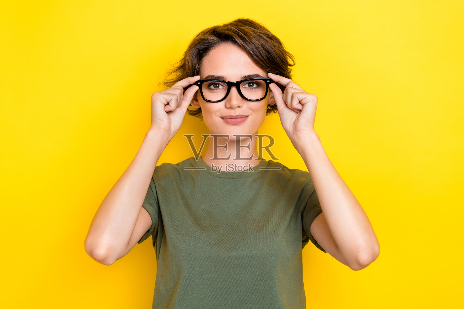 一位美丽迷人的短发女性，穿着卡其色T恤，双手触摸眼镜，背景为黄色。照片摄影图片
