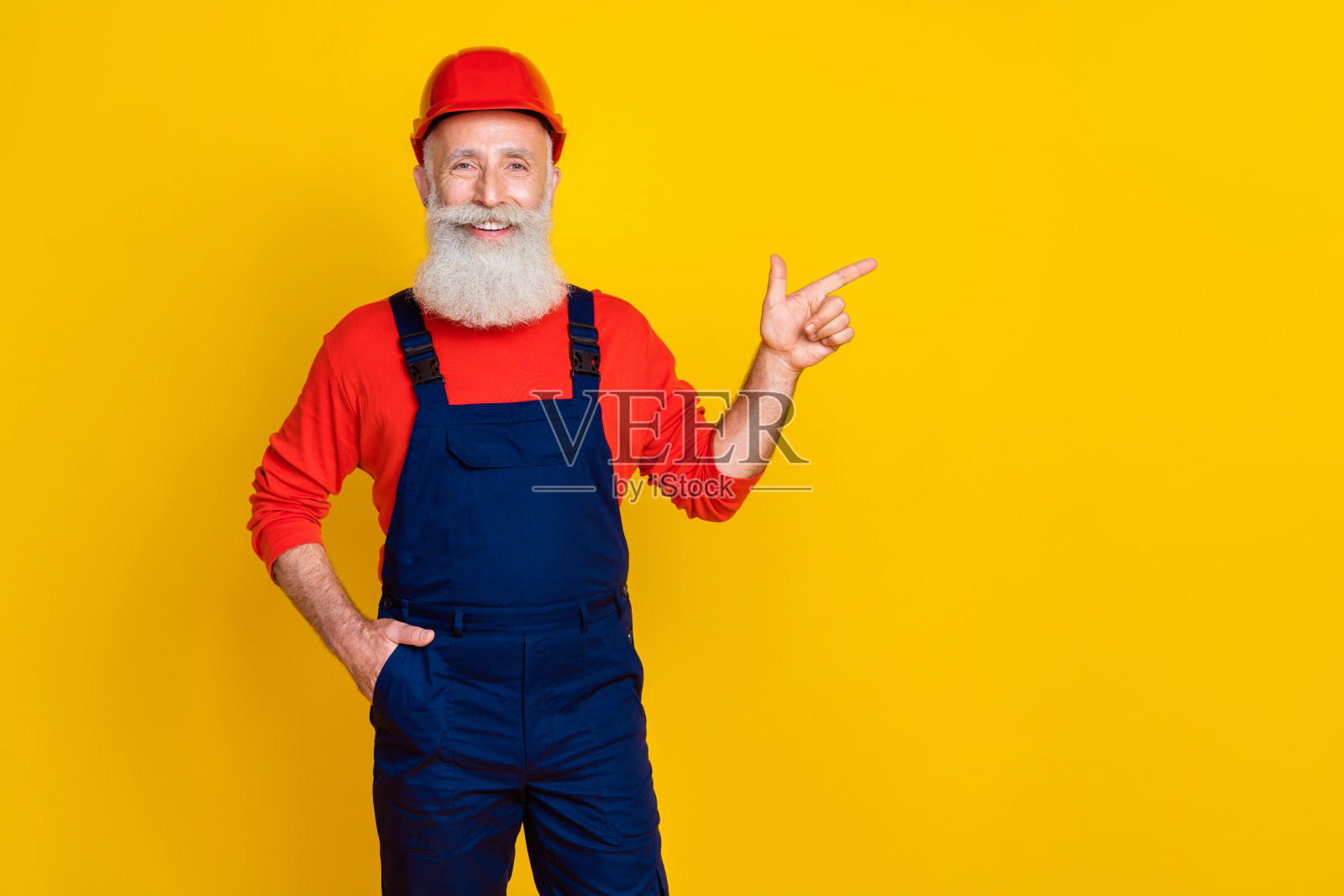 穿着红色工作服的快乐可爱老年人指着空白处，背景为黄色。照片摄影图片