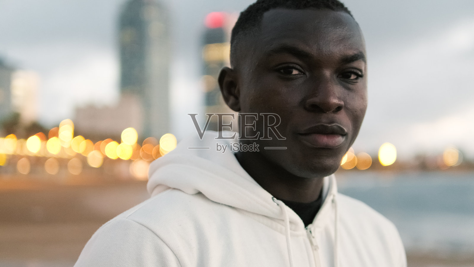 一位充满动力的运动员穿着白色连帽衫，在城市灯光和海洋的背景下为街头健身做准备，展现运动动力和积极的生活方式。照片摄影图片