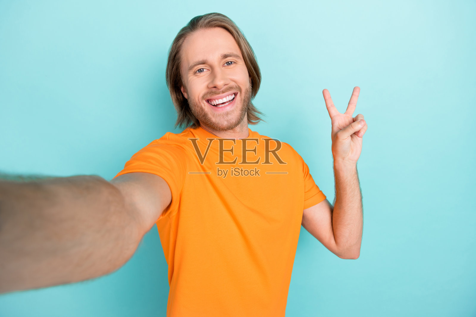 穿着橙色T恤的快乐积极男孩在青色背景下做出V字手势自拍照片摄影图片