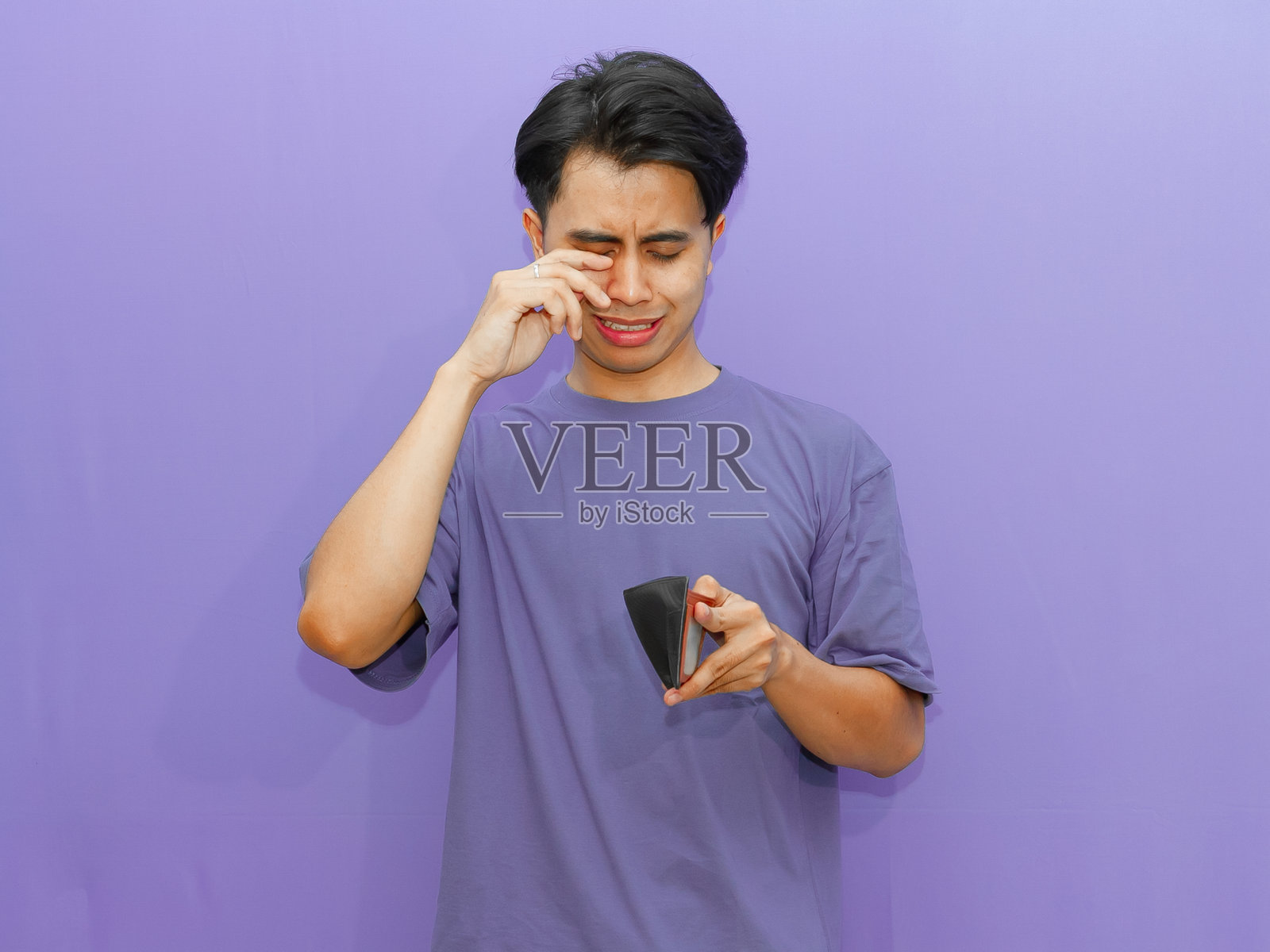 一位可怜的年轻亚洲男子手持空钱包，面露哭泣的表情，背景为紫色，突显经济困境。照片摄影图片