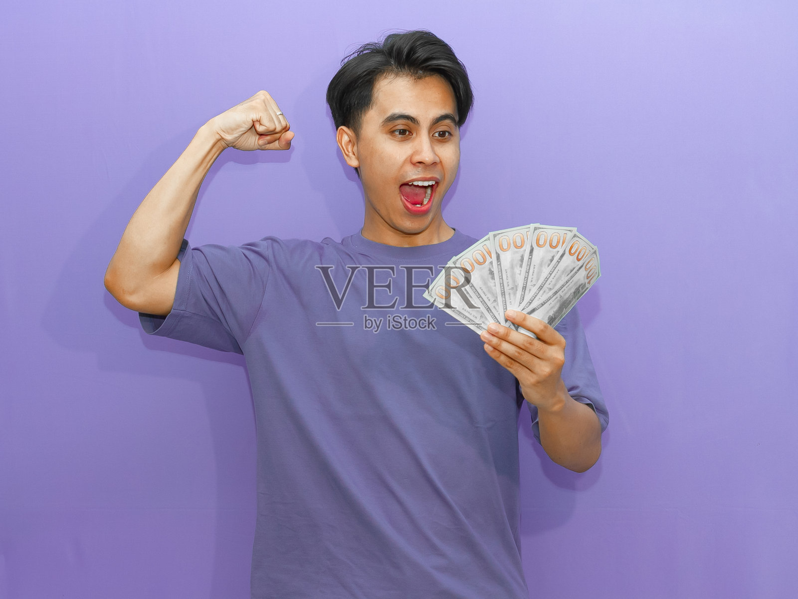 兴奋的年轻亚洲男子手握钞票，面带喜悦的表情庆祝成功，适合在充满活力的紫色背景上用于广告和促销。照片摄影图片