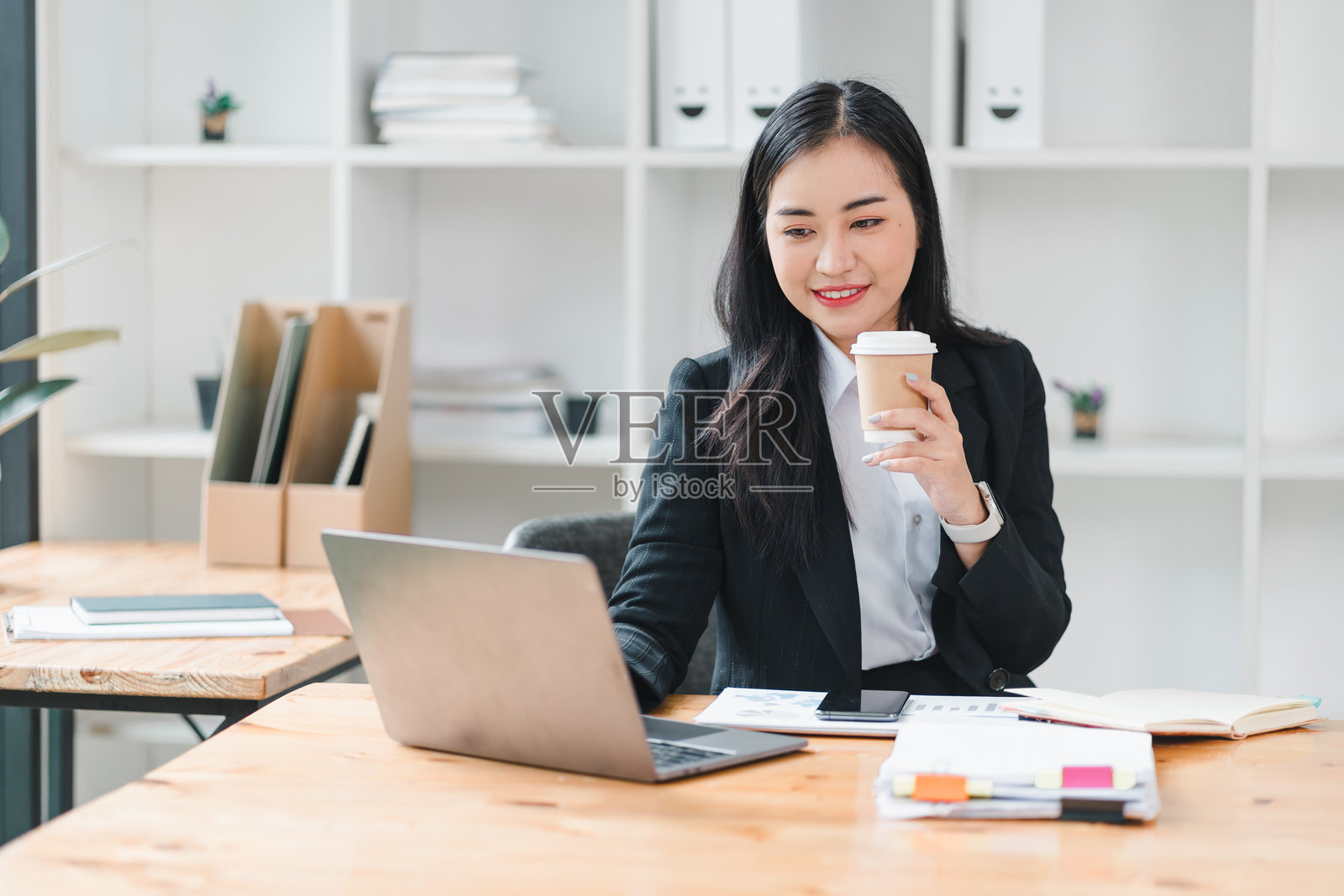 穿着黑色西装的职业女性坐在木桌前，微笑着手握咖啡杯，正在使用笔记本电脑。现代办公室环境明亮且井然有序，营造出高效的氛围。照片摄影图片