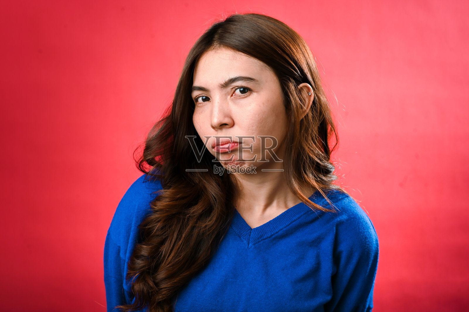 一位穿着蓝色毛衣的亚洲女性站在红色背景前，脸上带着顽皮的撅嘴，散发出轻松愉快和微妙幸福的气息。照片摄影图片