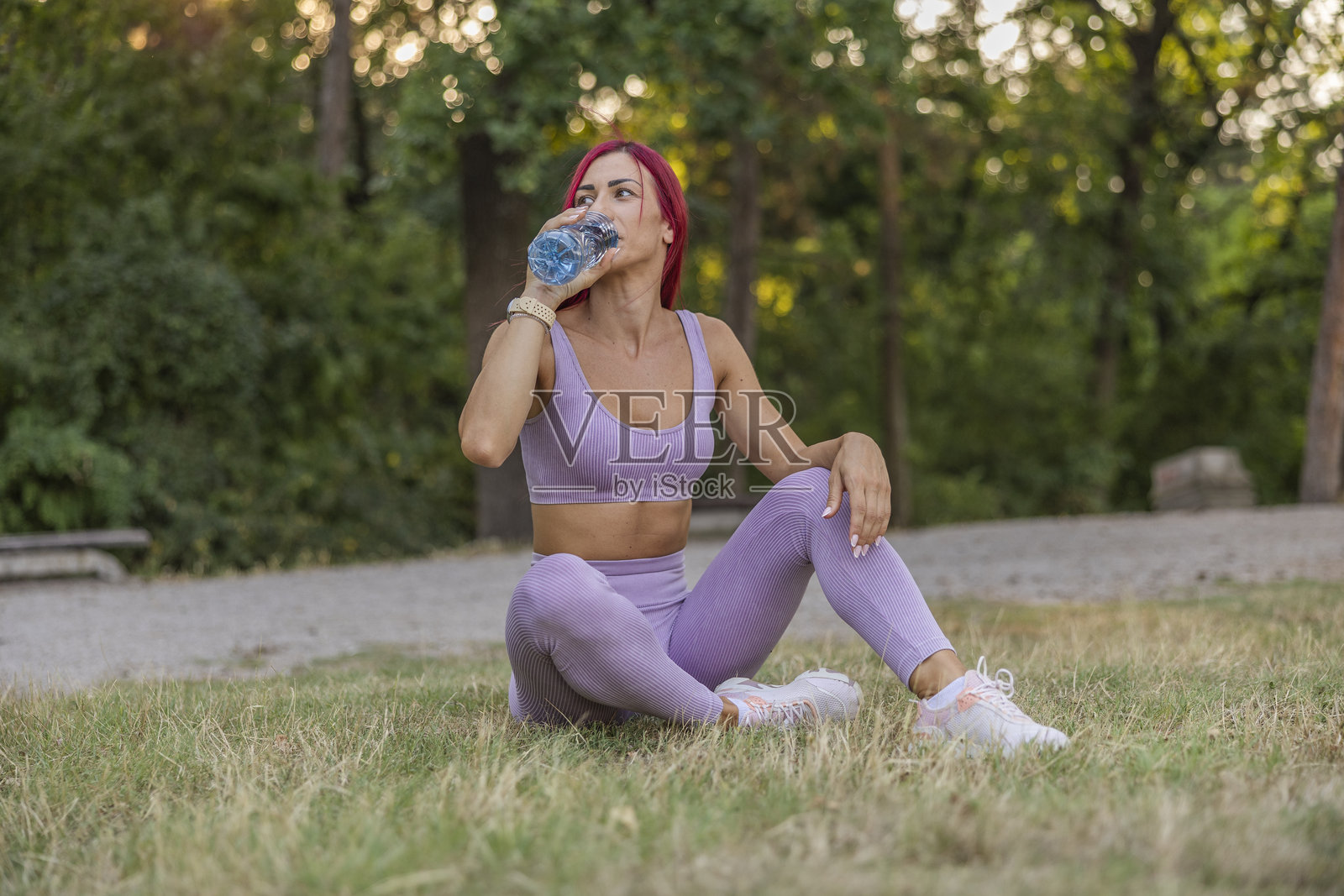 穿着运动服的女性在森林中坐着，喝着瓶装新鲜水，享受户外慢跑的乐趣。照片摄影图片