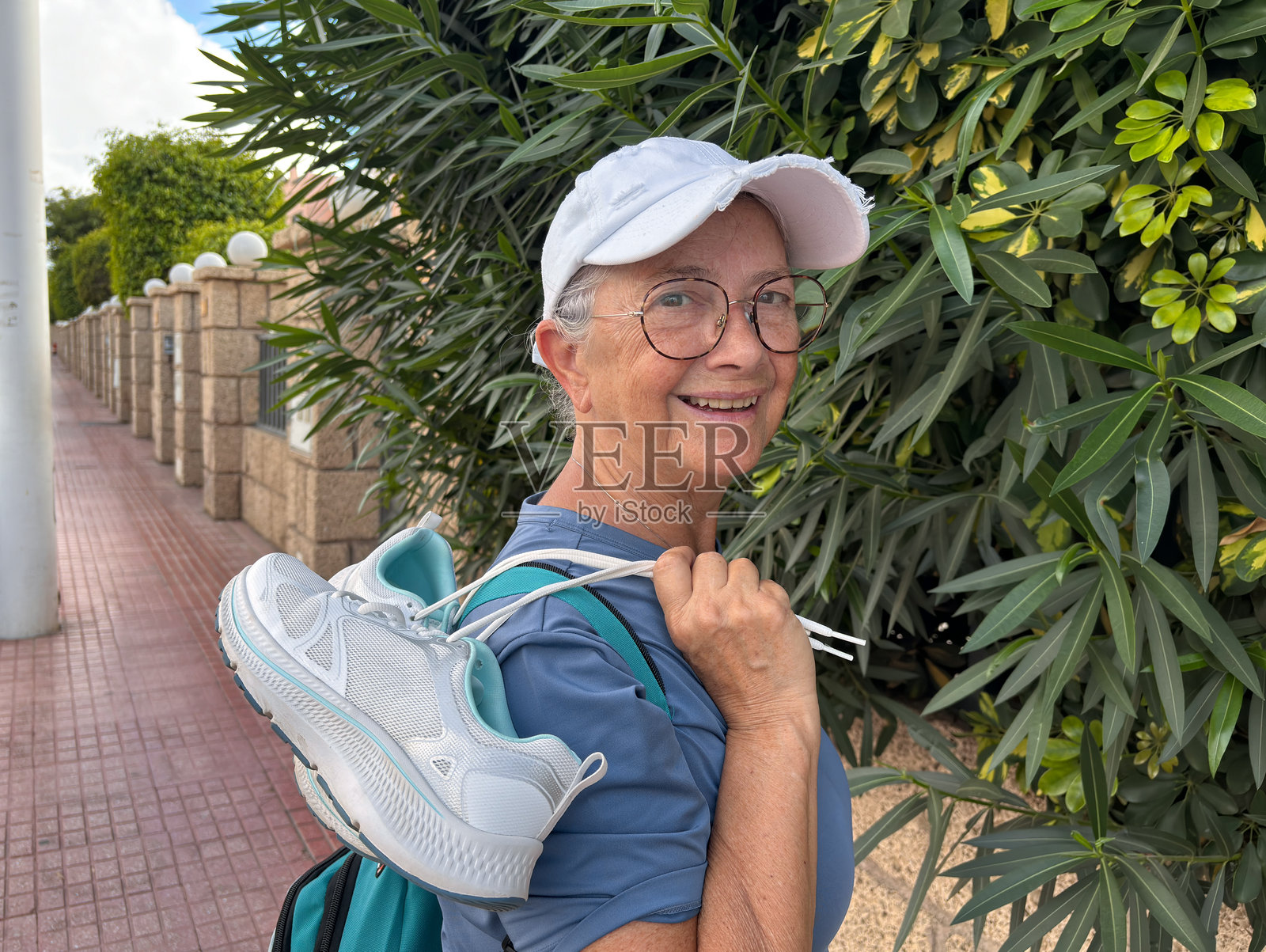 一位戴着帽子和眼镜的活跃老年女性在街上走着，因脚疼而将鞋子扛在肩上，正看着镜头。照片摄影图片