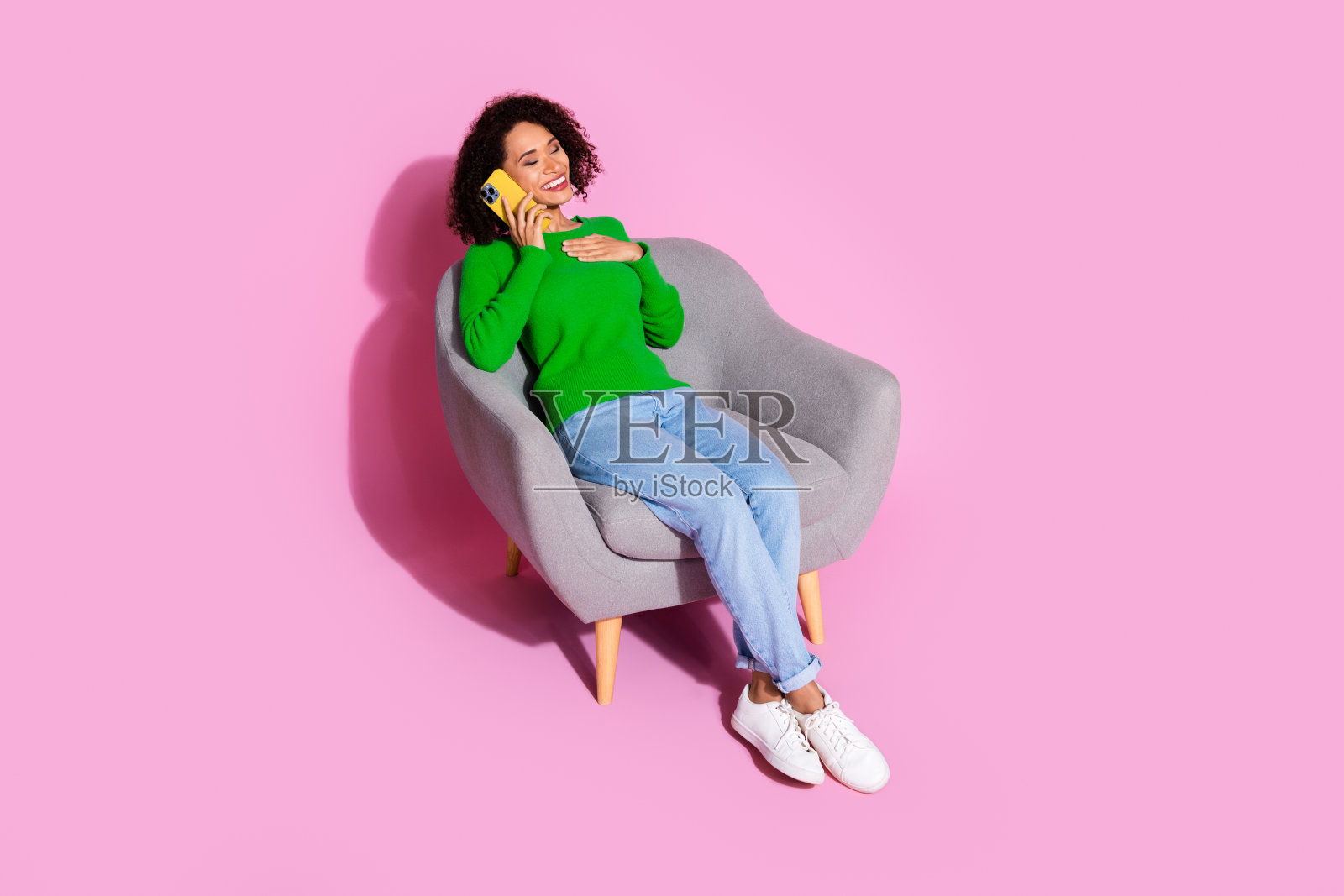 全身照，吸引人的年轻女性坐在柔软的扶手椅上，穿着时尚的绿色衣服，打电话，背景为粉色。照片摄影图片