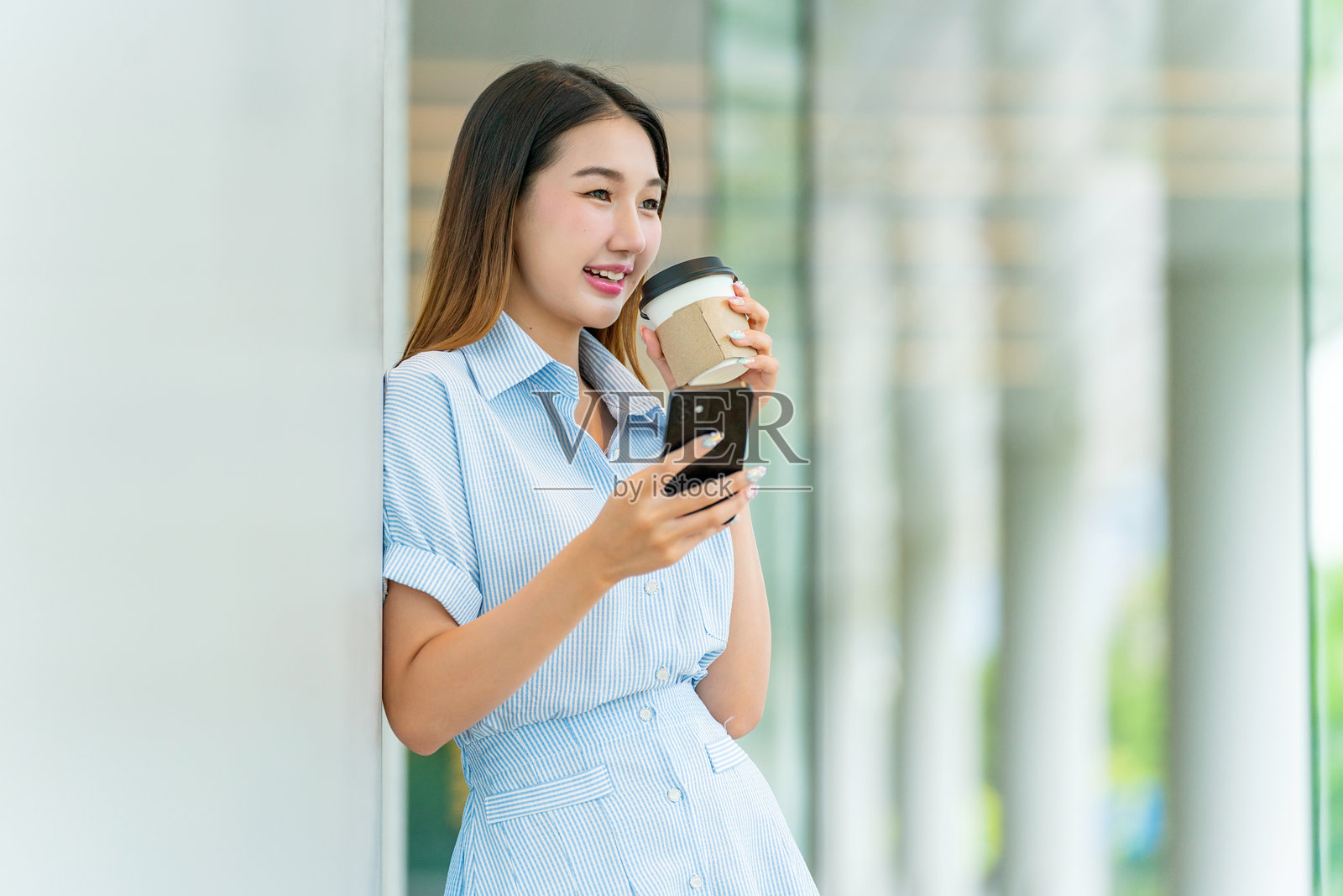 年轻亚洲女性在玻璃窗前享用咖啡，同时使用手机，窗外映射出城市景观。照片摄影图片