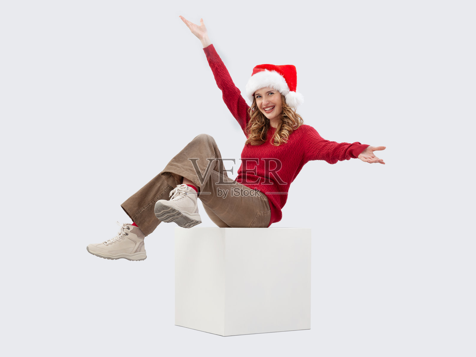 圣诞快乐！美丽快乐的年轻金发女性坐在白色立方体上，顽皮地张开双臂，面向镜头。她戴着圣诞老人帽，背景为白色。照片摄影图片