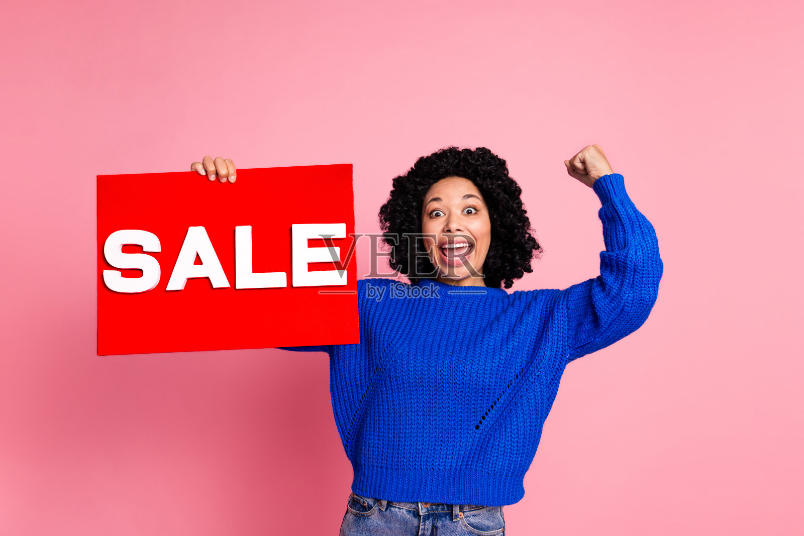 穿着蓝色针织毛衣的搞笑年轻女性，手握红色促销海报，拳头举起，背景为粉色。照片摄影图片