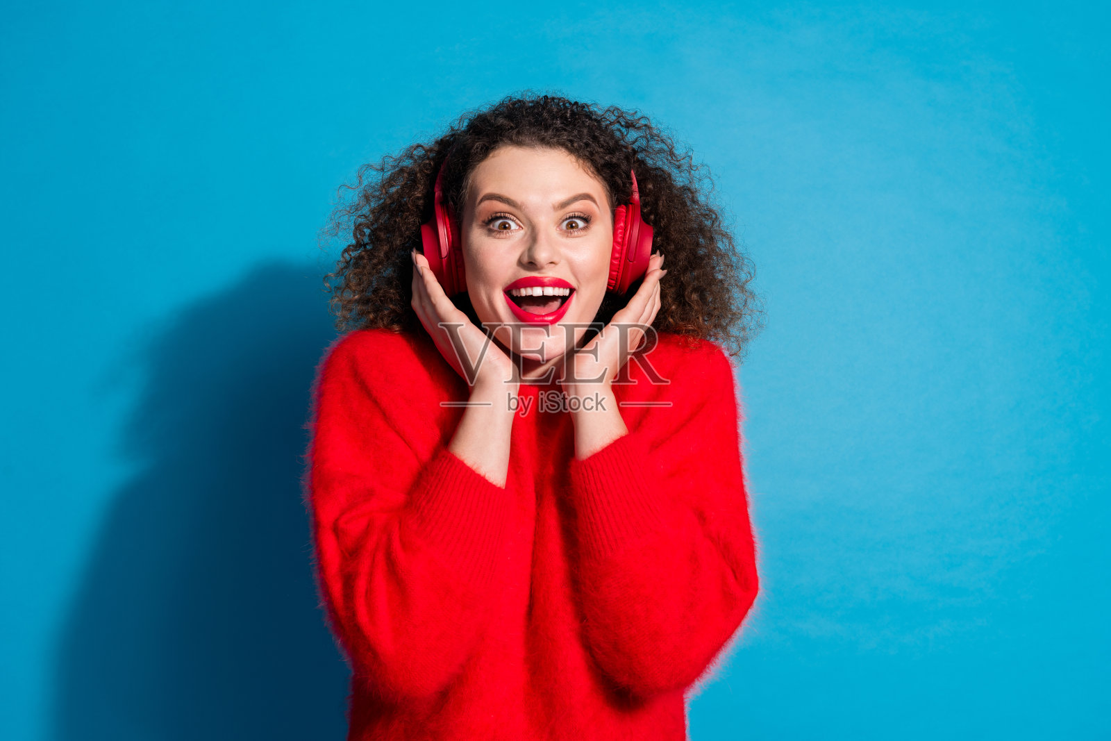 漂亮年轻女孩佩戴耳机兴奋地听音乐，穿着时尚的红色服装，背景为蓝色。照片摄影图片