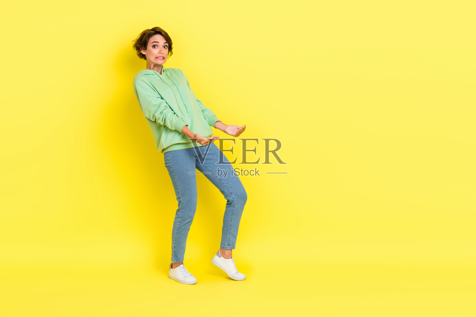 穿着绿色连帽衫的惊讶时尚女性全身照，双臂张开，空旷的空间，背景为黄色。照片摄影图片