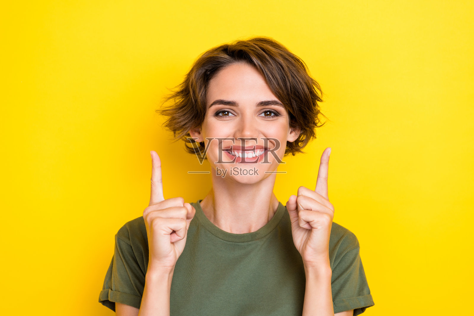 漂亮女孩穿着卡其色T恤，留着波波头，指向空白区域，背景为黄色，适合购物主题。照片摄影图片