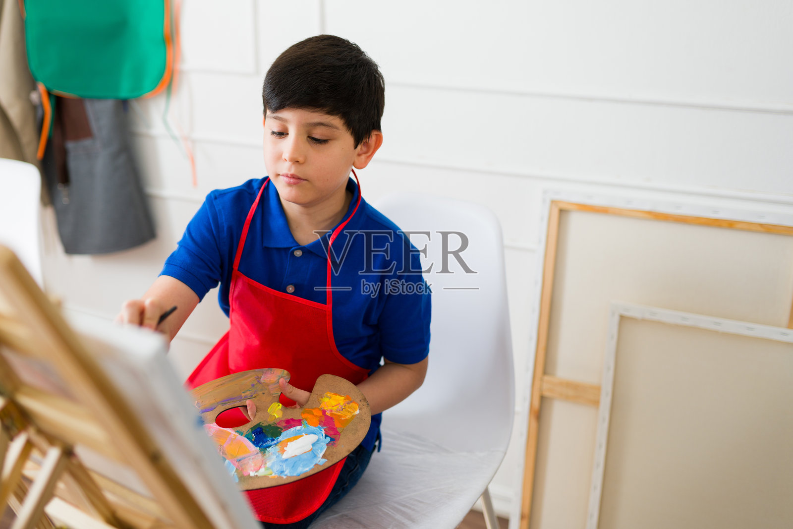 孩子穿着红色围裙，在美术课上用小画架作画，手里拿着五彩斑斓的调色板。照片摄影图片