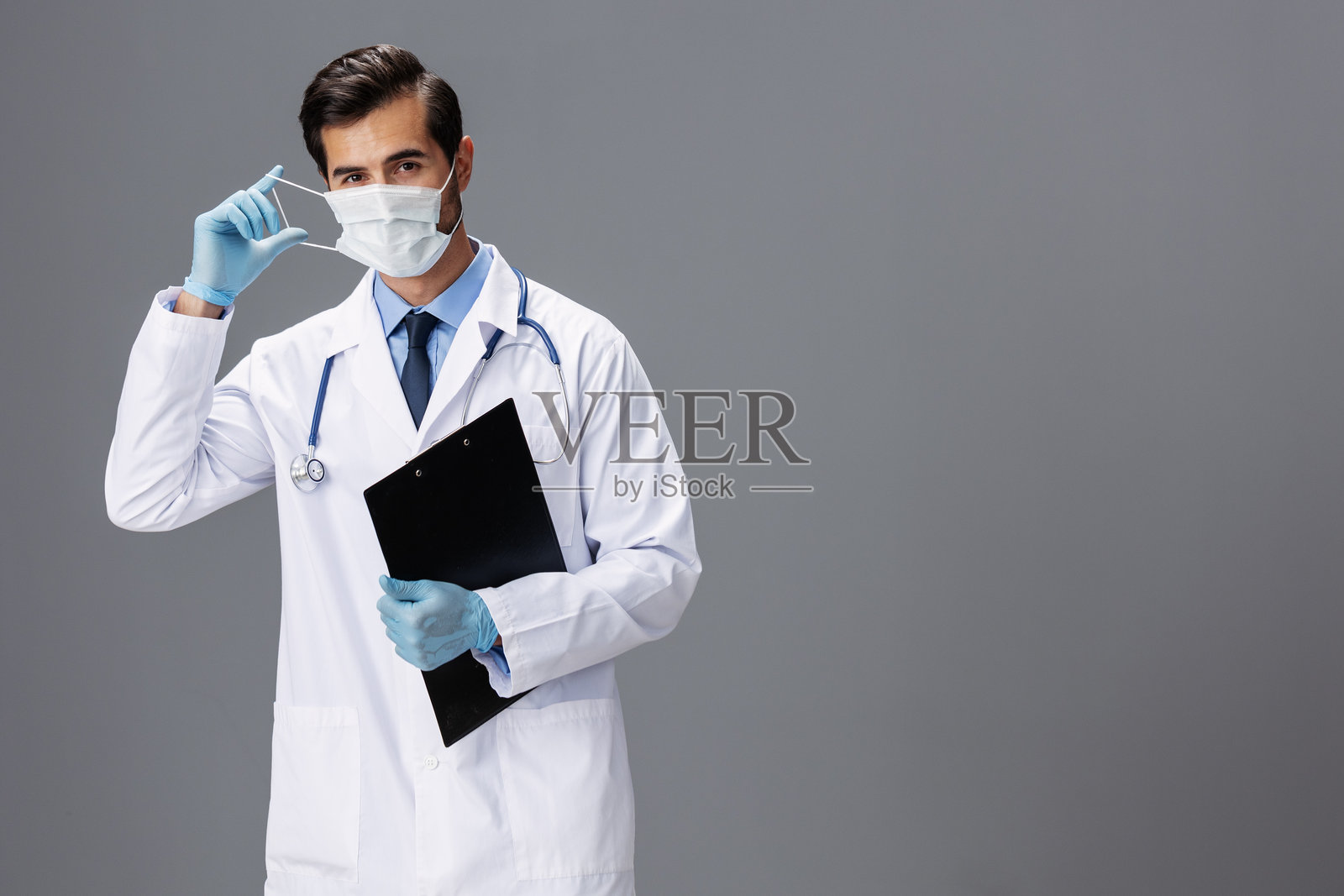 一位穿着白大褂和医用口罩的男性医生手持笔记本，面向镜头，背景为灰色，留有空白区域，适合添加文字。照片摄影图片