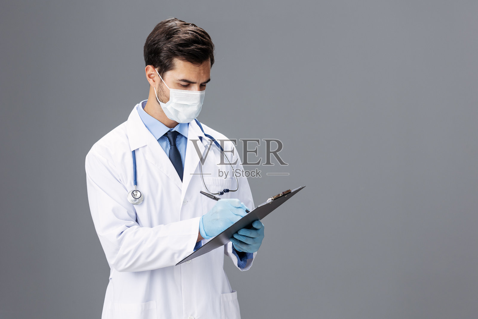 一位穿着白大褂和医用口罩的男性医生，手中拿着记事本和病历卡，正对着镜头，背景为灰色，留有空白区域，适合添加文字。照片摄影图片