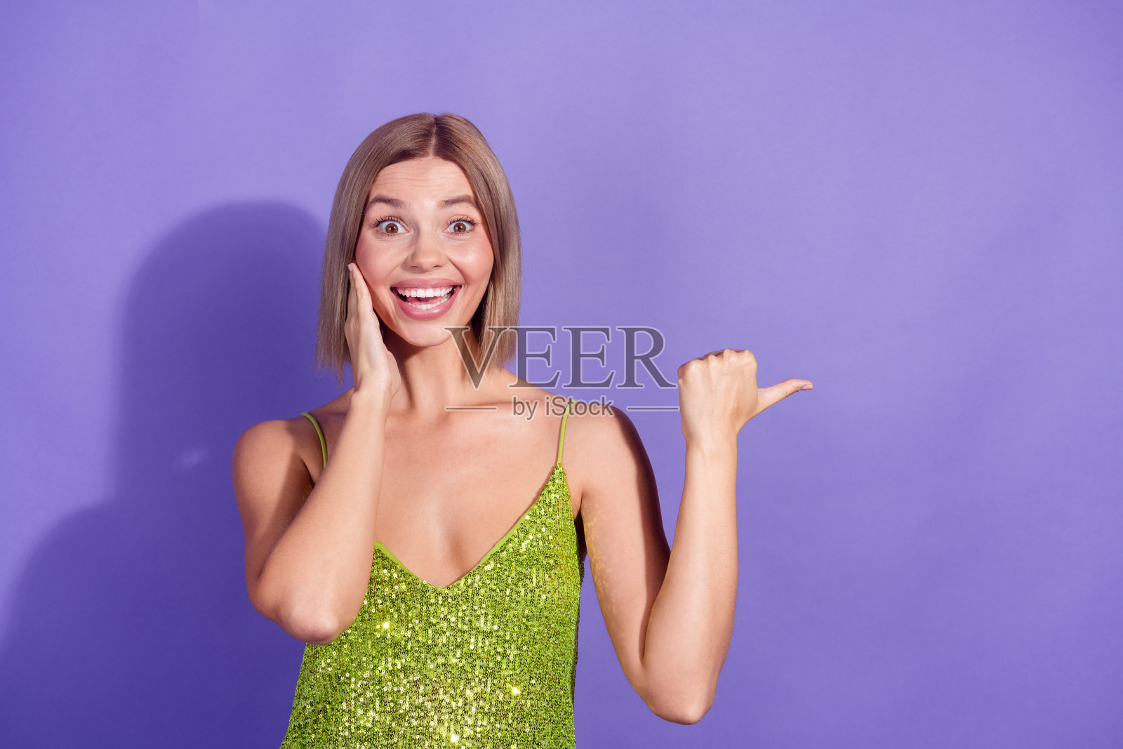 金发年轻女性穿着绿色闪亮背心，指着空白处，惊讶地表达，背景为紫色。照片摄影图片