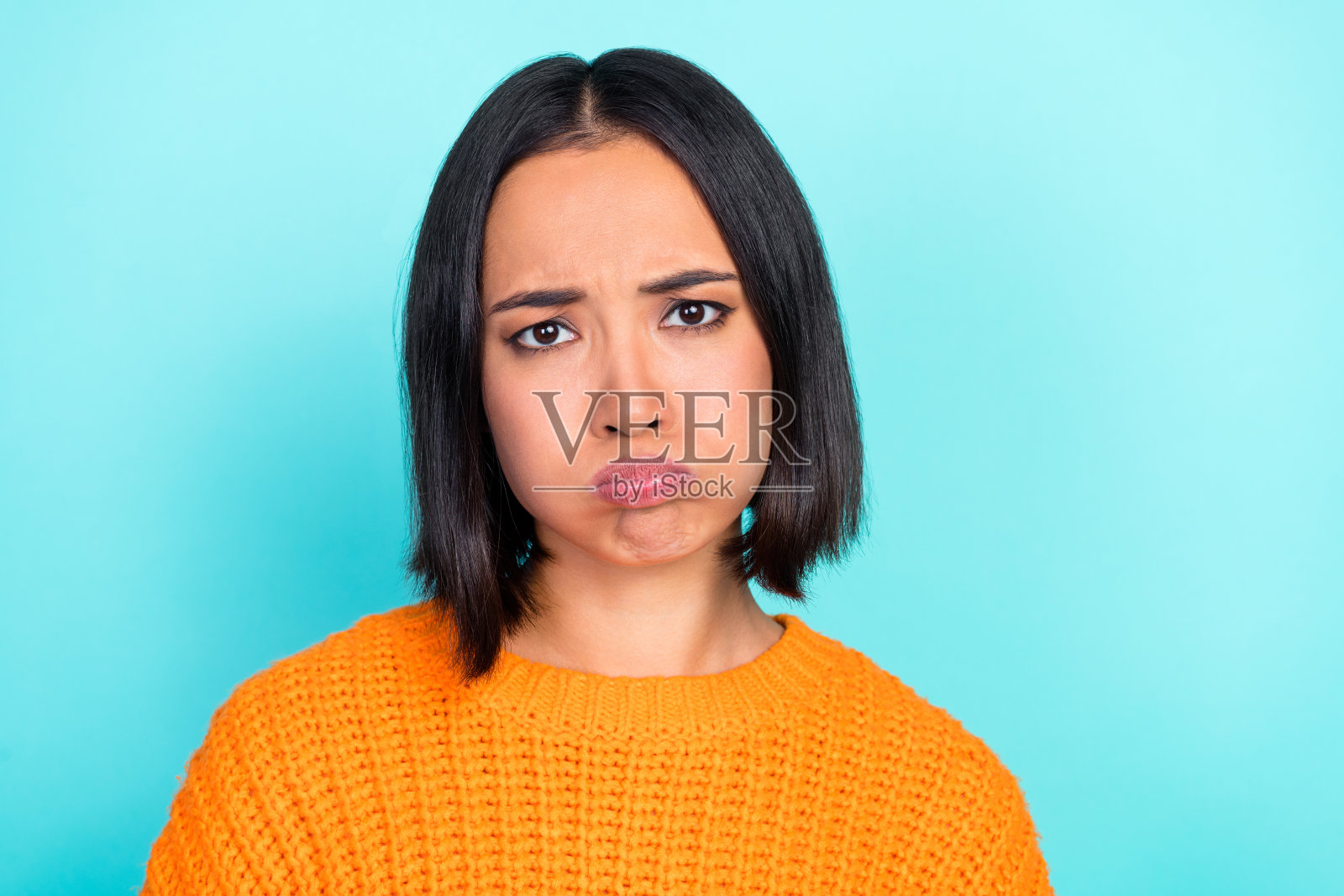 一位可爱的小女孩，直发，穿着橙色毛衣，嘴唇丰满，表情悲伤，孤立在青色背景上。照片摄影图片