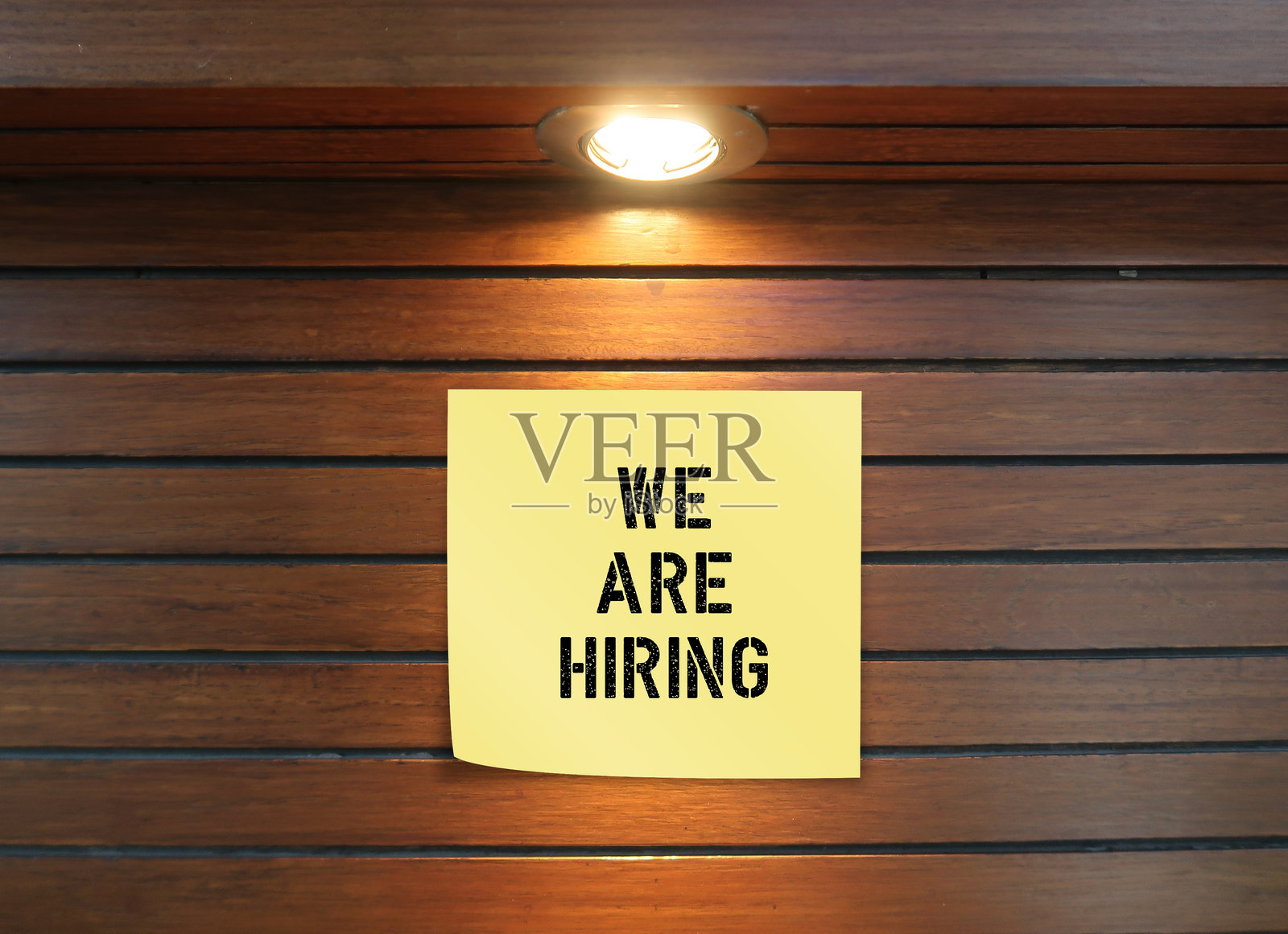 黄色便条上写着“我们在招聘”，贴在木质背景上，顶部有灯光，象征着企业扩展，寻找员工或工人。照片摄影图片