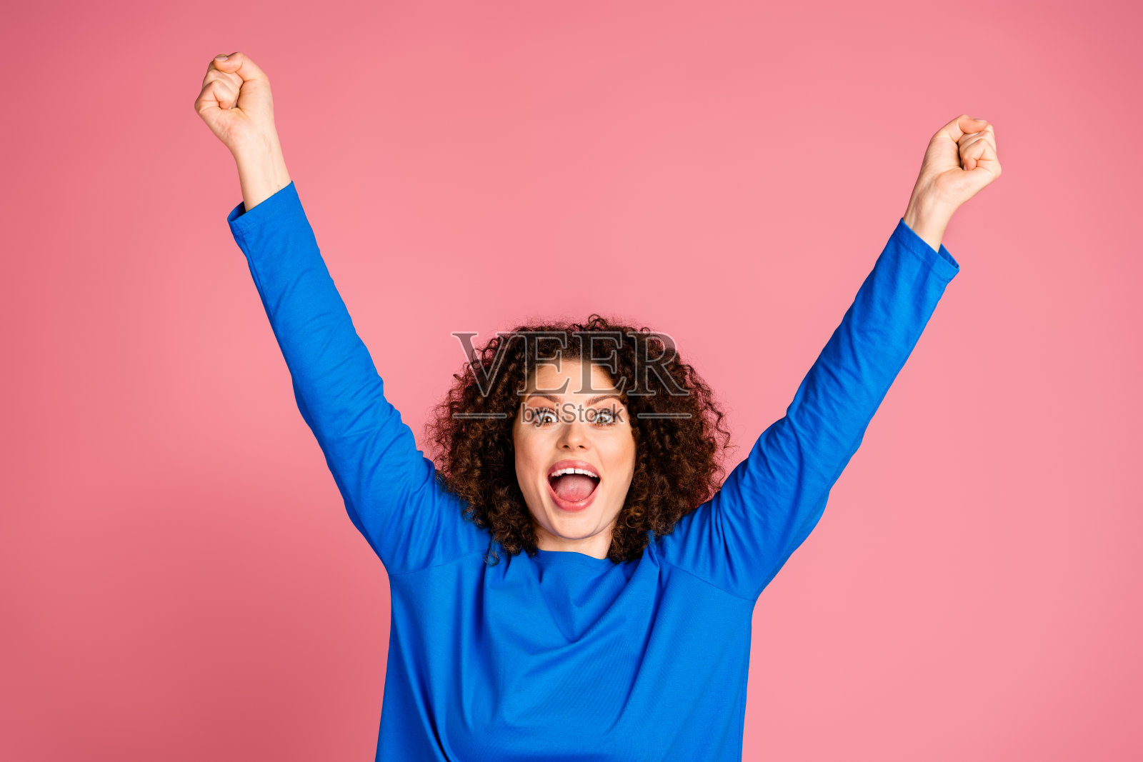 兴奋的年轻女性穿着休闲蓝色毛衣，在粉色背景前举起双臂庆祝照片摄影图片