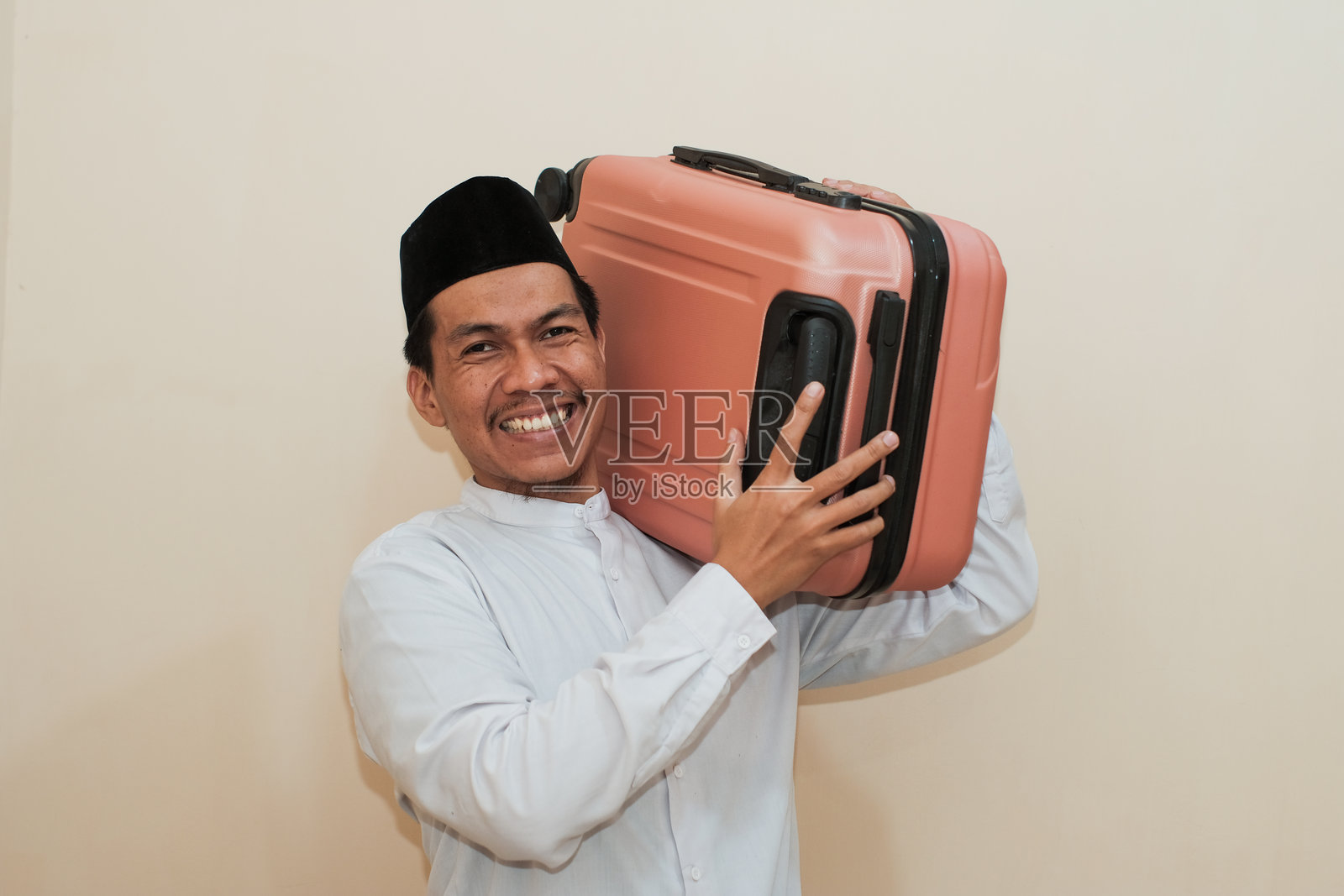 微笑的东南亚穆斯林男子手提行李箱，脸上洋溢着庆祝开斋节的快乐。背景为白色。照片摄影图片