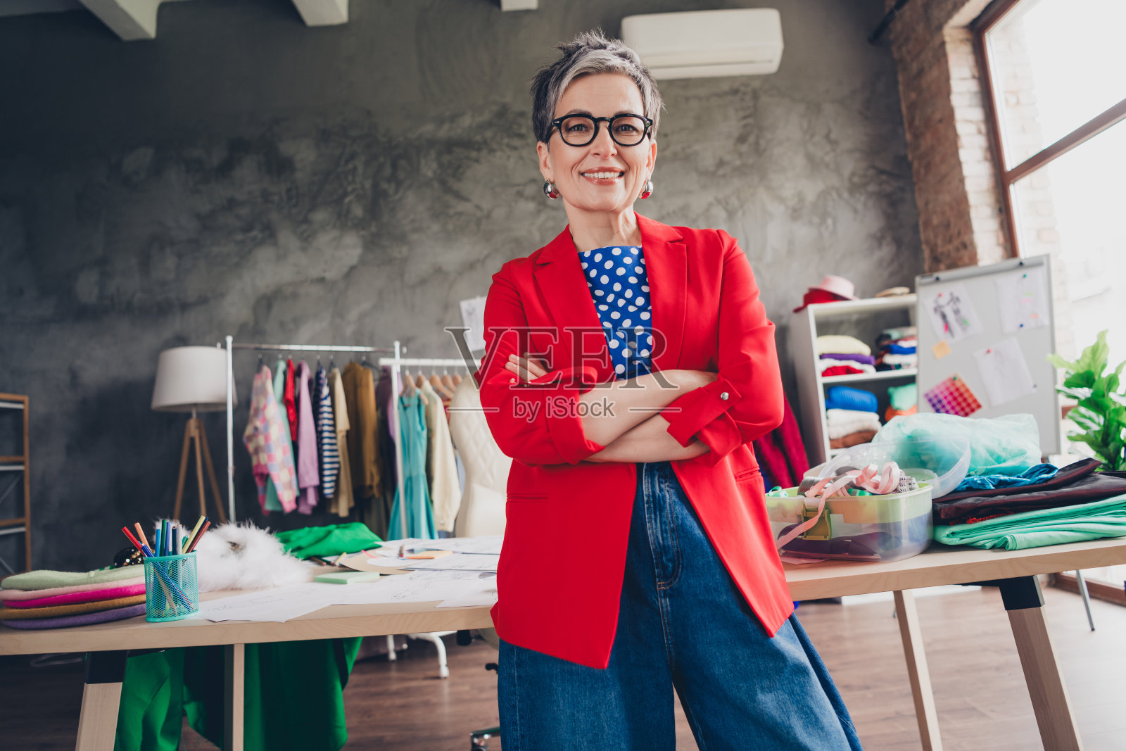 穿着时尚的女裁缝设计师，双臂交叉，坐在室内的阁楼办公室工作室里照片摄影图片
