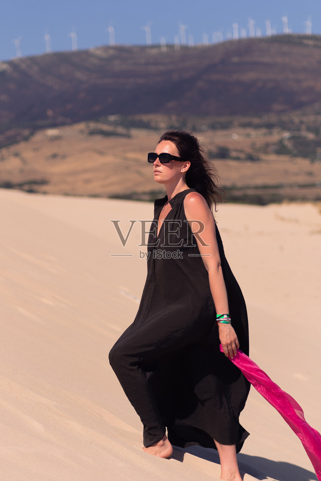 穿着长黑裙、佩戴粉色围巾和太阳镜的美丽女孩在沙丘上摆姿势。阳光明媚的夏日，壮丽的大沙丘，女人独自在沙漠中。照片摄影图片