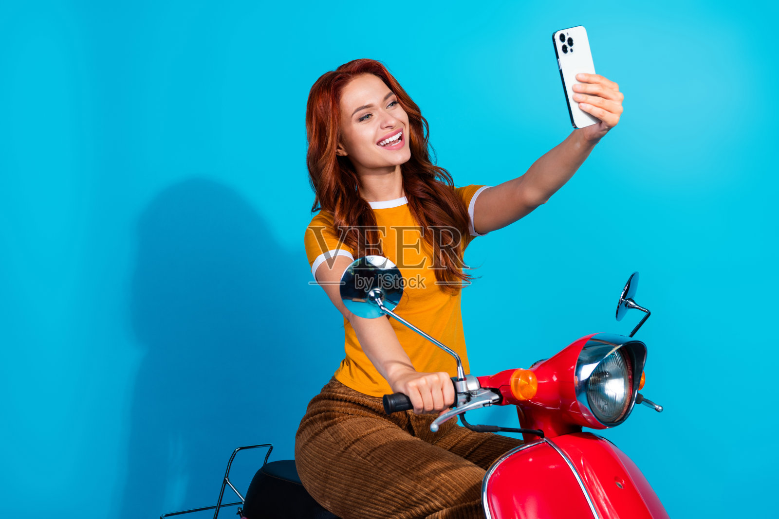 穿着时尚的可爱快乐女性骑着摩托车，手持电子设备，背景为蓝色。照片摄影图片