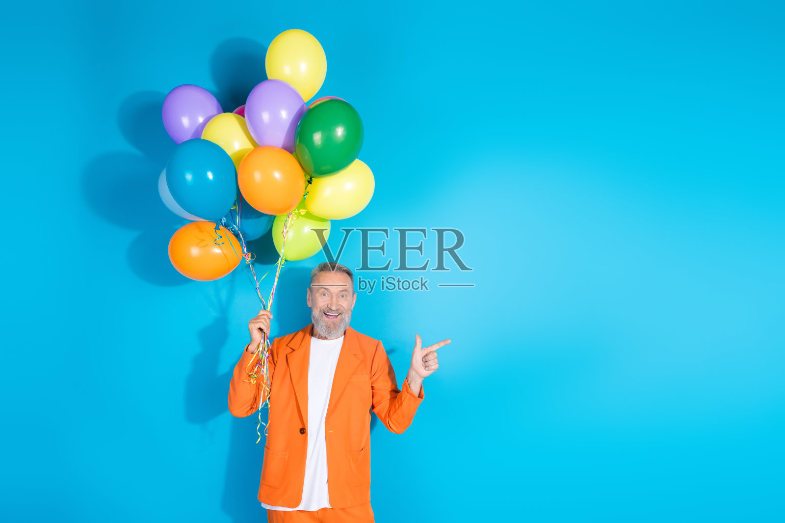 时尚的中年男子穿着橙色服装，手指指向空旷的地方，背景为蓝色，手中拿着气球。照片摄影图片