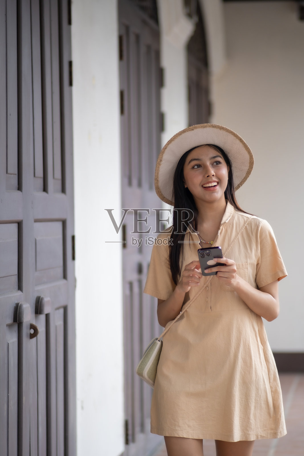 一位戴着帽子、穿着棕褐色连衣裙的女性正在拿着手机。照片摄影图片