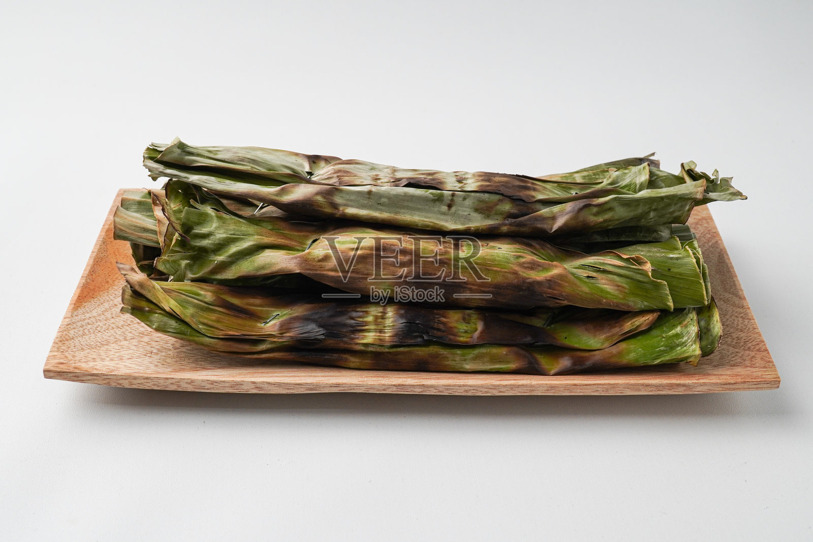 Otak-otak是一道用剁碎的鲭鱼包裹在香蕉叶中，烤制而成的菜肴，通常配有花生酱。白色背景下的木盘上的Otak-otak，拍摄角度为30度。照片摄影图片