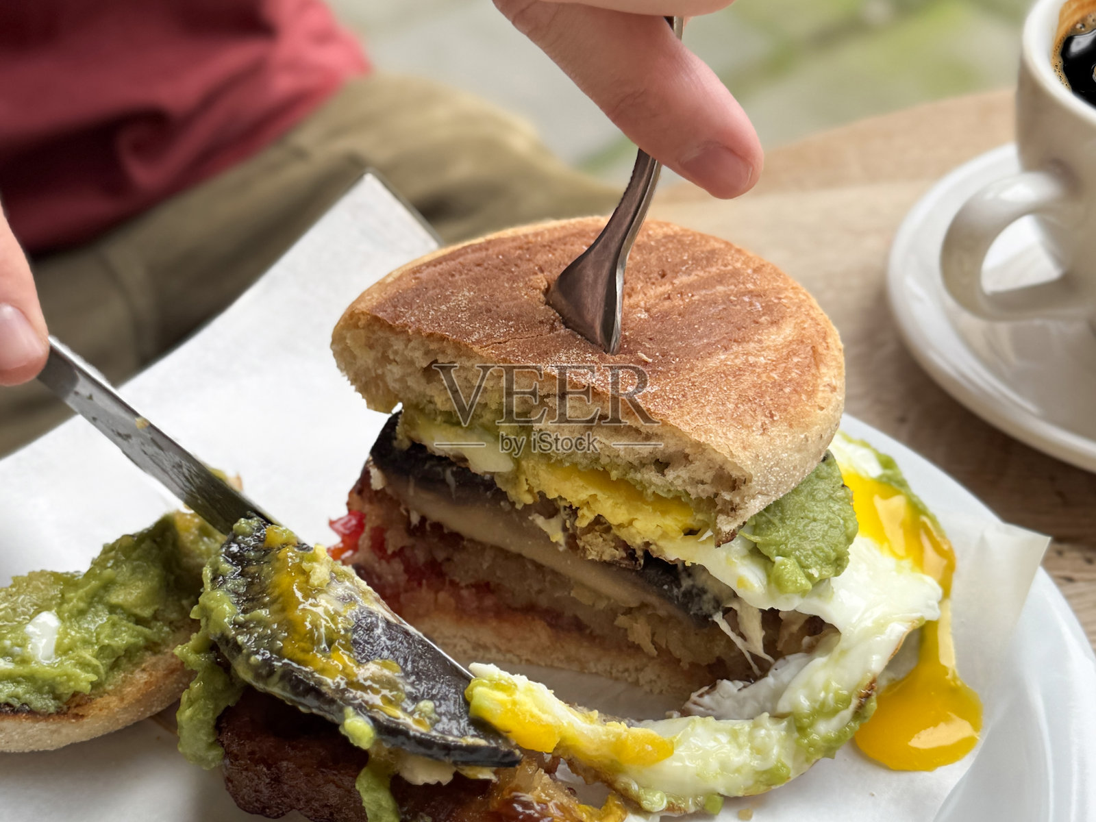 一位无法辨认的男子正在切割早餐，盘子里有吐司面包夹着辣椒果酱、香肠饼、波特贝罗蘑菇、煎蛋和捣碎的鳄梨，旁边是一杯咖啡，餐厅环境，重点在前景。照片摄影图片