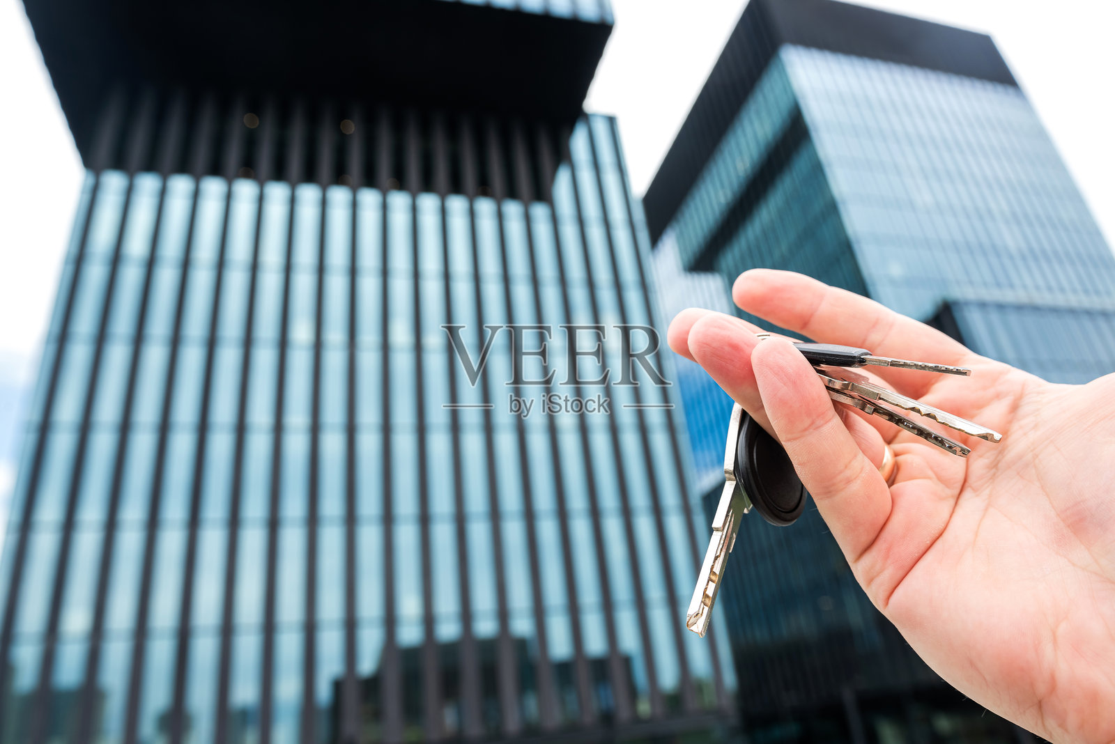 房地产经纪人手中的房屋钥匙与新建的摩天大楼玻璃办公室。购房或租房的概念。照片摄影图片