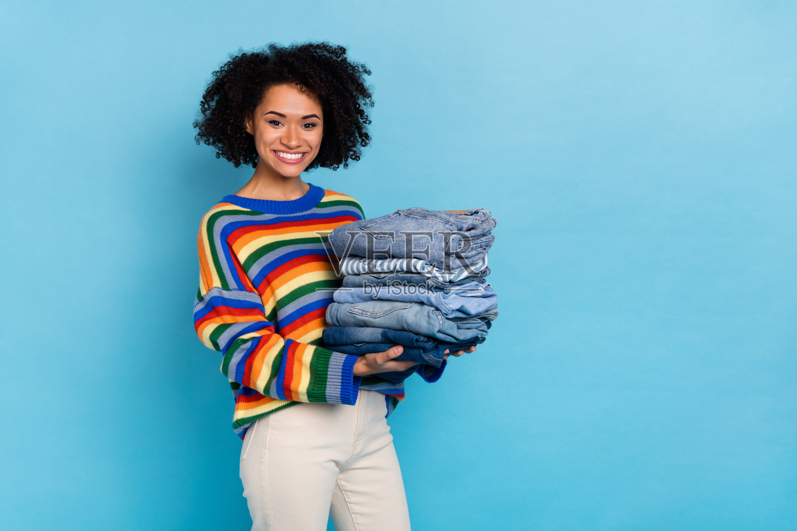 年轻短发女士手持牛仔裤，穿着彩色毛衣，背景为蓝色。照片摄影图片