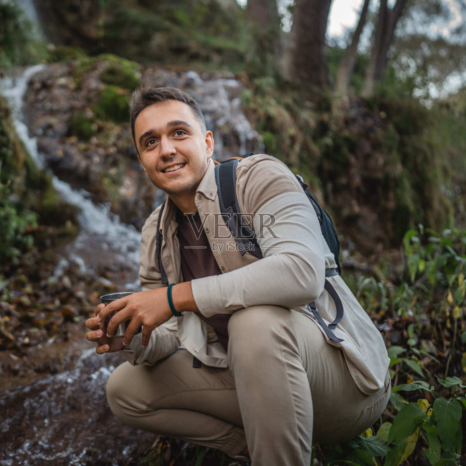 成年男性徒步旅行者蹲在河边，正在用水杯装水。照片摄影图片