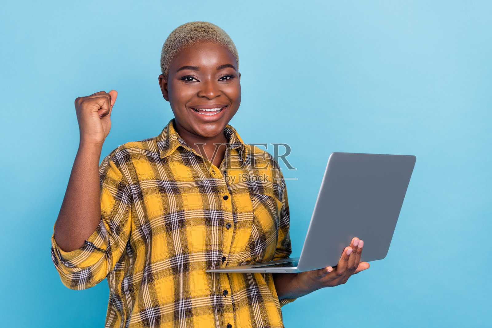 穿着格子衬衫的幸运漂亮女人举起拳头，手持现代设备，背景为蓝色。照片摄影图片