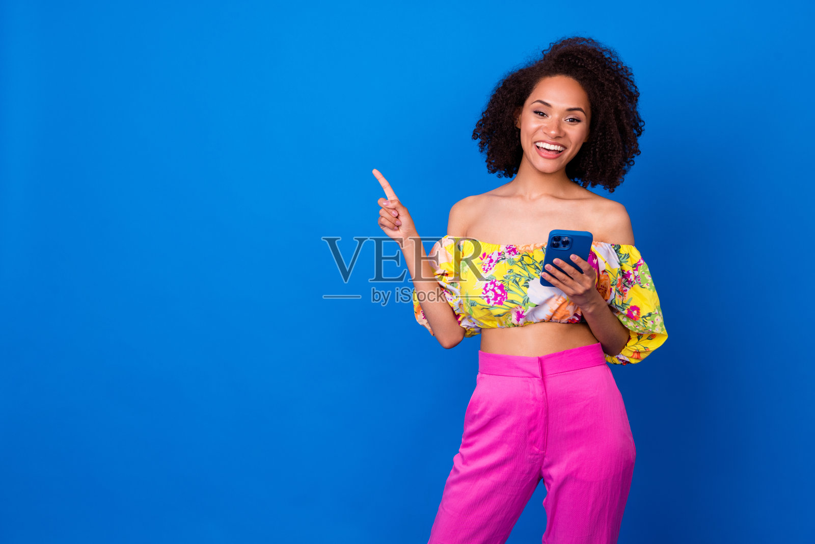 一位迷人的年轻女性，波浪卷发，手持设备，指向空白处，穿着时尚多彩的服装，背景为闪亮的蓝色。照片摄影图片
