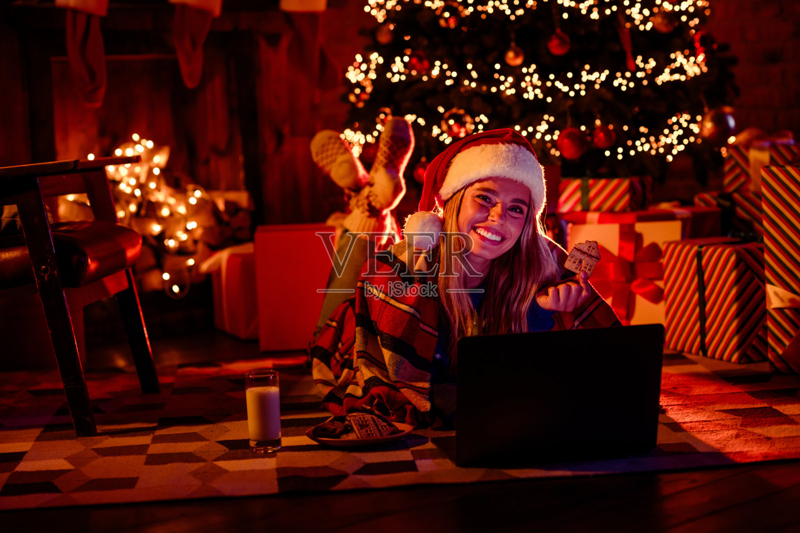 年轻女性穿着圣诞服装，坐在室内房间里观看电影，吃着饼干，周围装饰着花环和灯光，营造出节日气氛。照片摄影图片