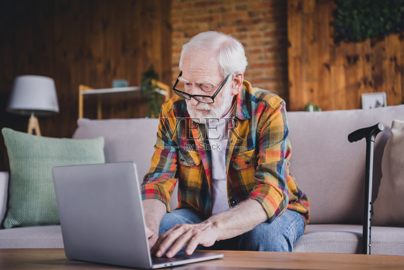 自信聪明的退休男士穿着格子衬衫，戴着眼镜，坐在沙发上使用现代设备，室内公寓环境。照片摄影图片