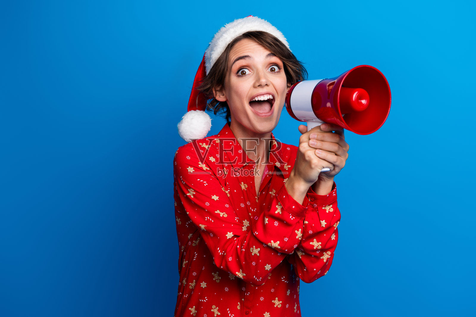 穿着红色圣诞新年衬衫的可爱年轻女孩，手持扬声器，背景为空白的蓝色。照片摄影图片