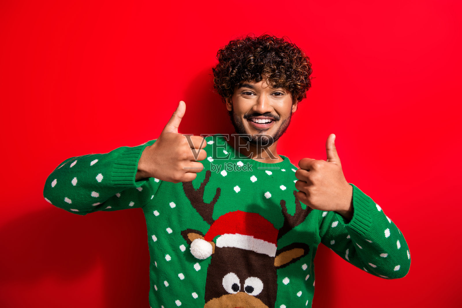 年轻男子穿着新年圣诞毛衣，竖起大拇指，背景为红色。照片摄影图片