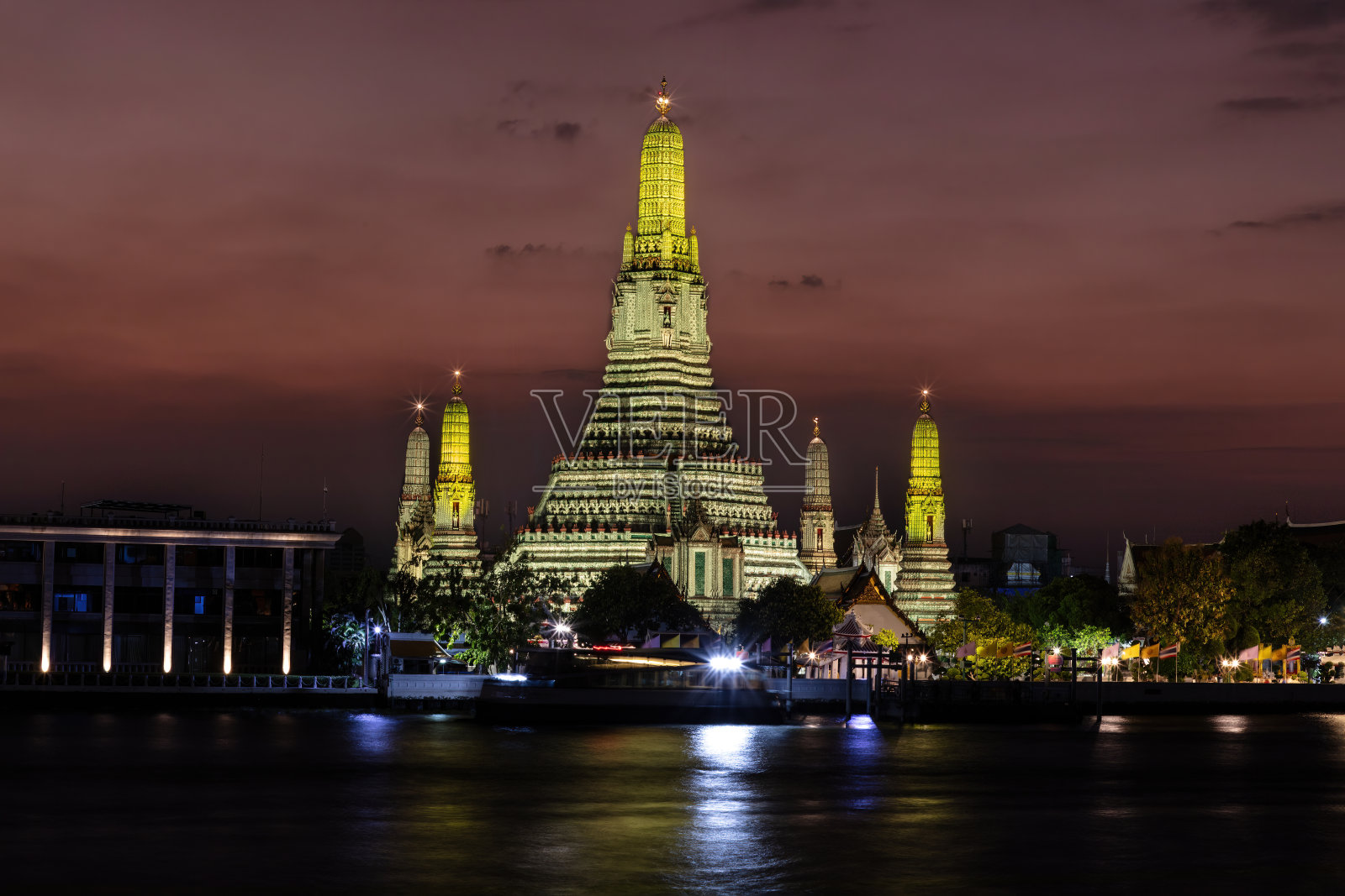 曼谷的黎明寺在黄昏时分。寺庙和尖塔沐浴在黄色的灯光中。深橙色的天空。前景是河流。照片摄影图片