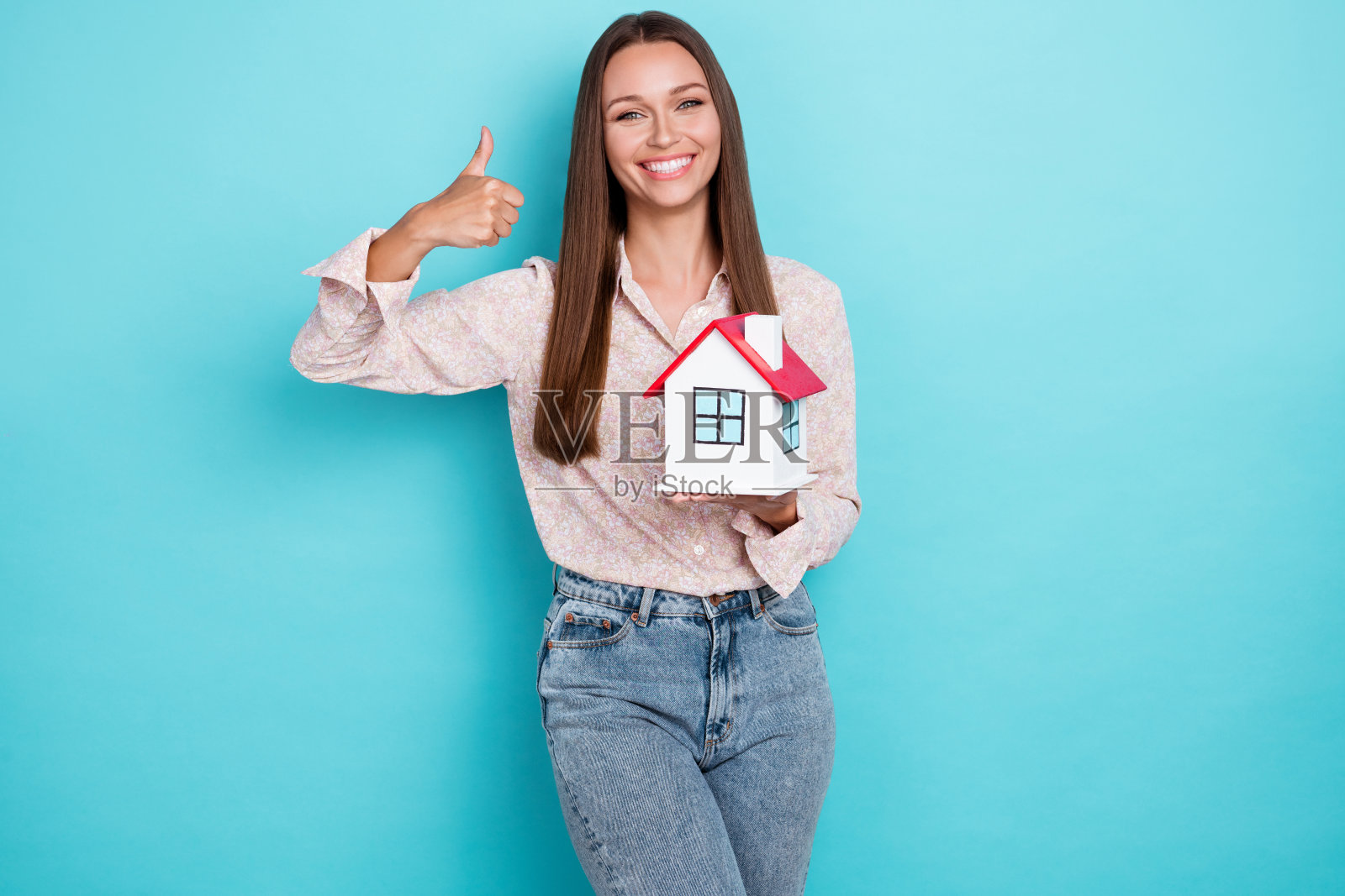 时尚的千禧女性穿着白衬衫和牛仔裤，竖起大拇指，手持房屋，背景为蓝色。照片摄影图片
