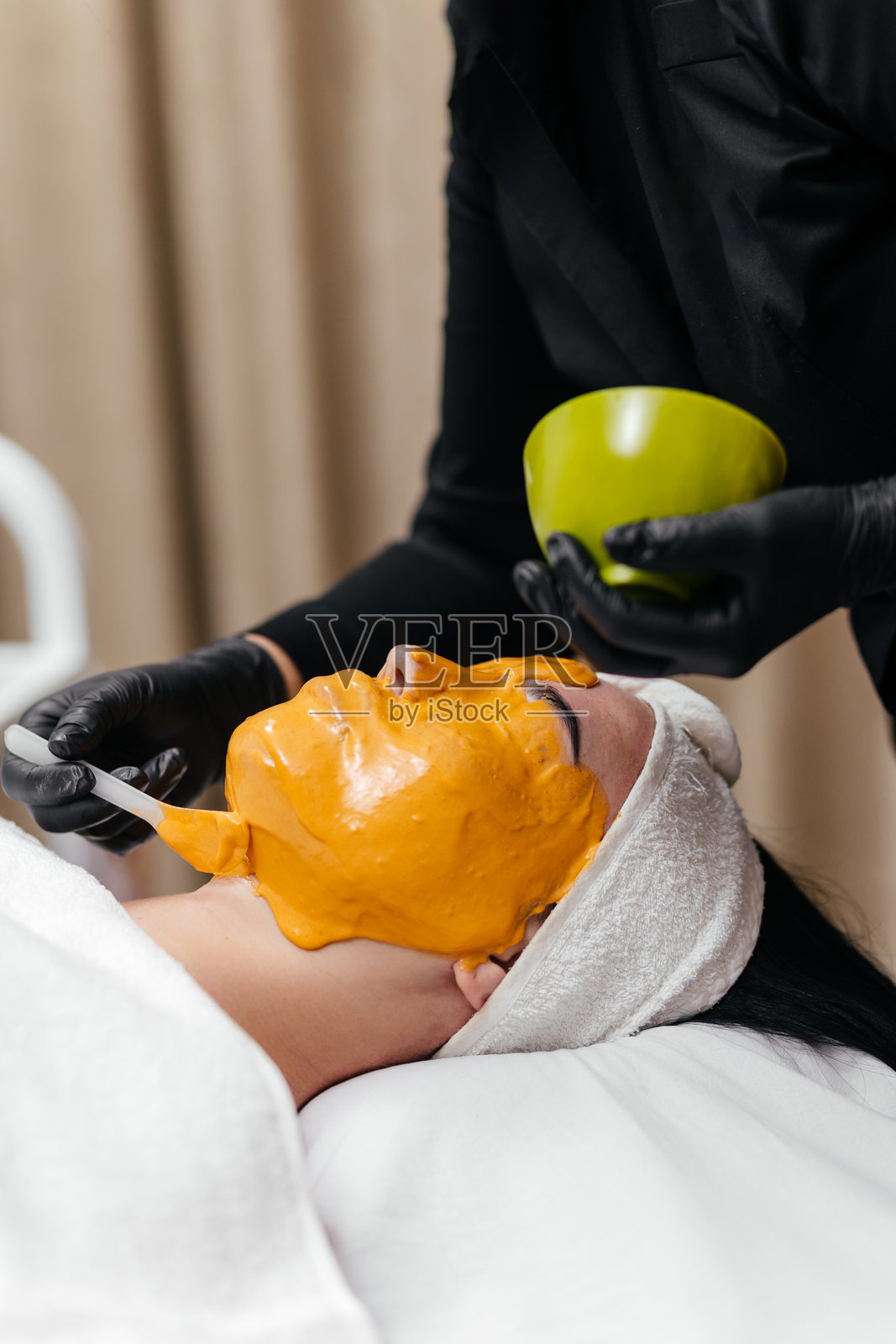 美容师用刮刀将藻酸盐面膜涂抹在客户脸上的特写镜头。场景突显了美容院专业的面部护肤疗程。照片摄影图片